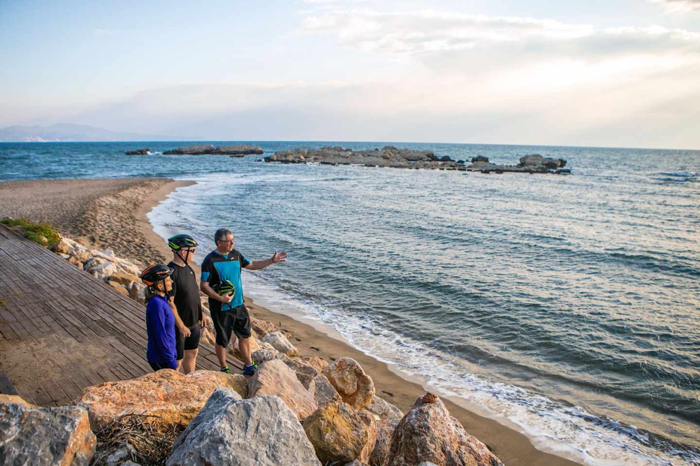 A wooden walkway along the rocky coastline, with two people standing and enjoying the view of the serene ocean and distant rocky formations under a partly cloudy sky.