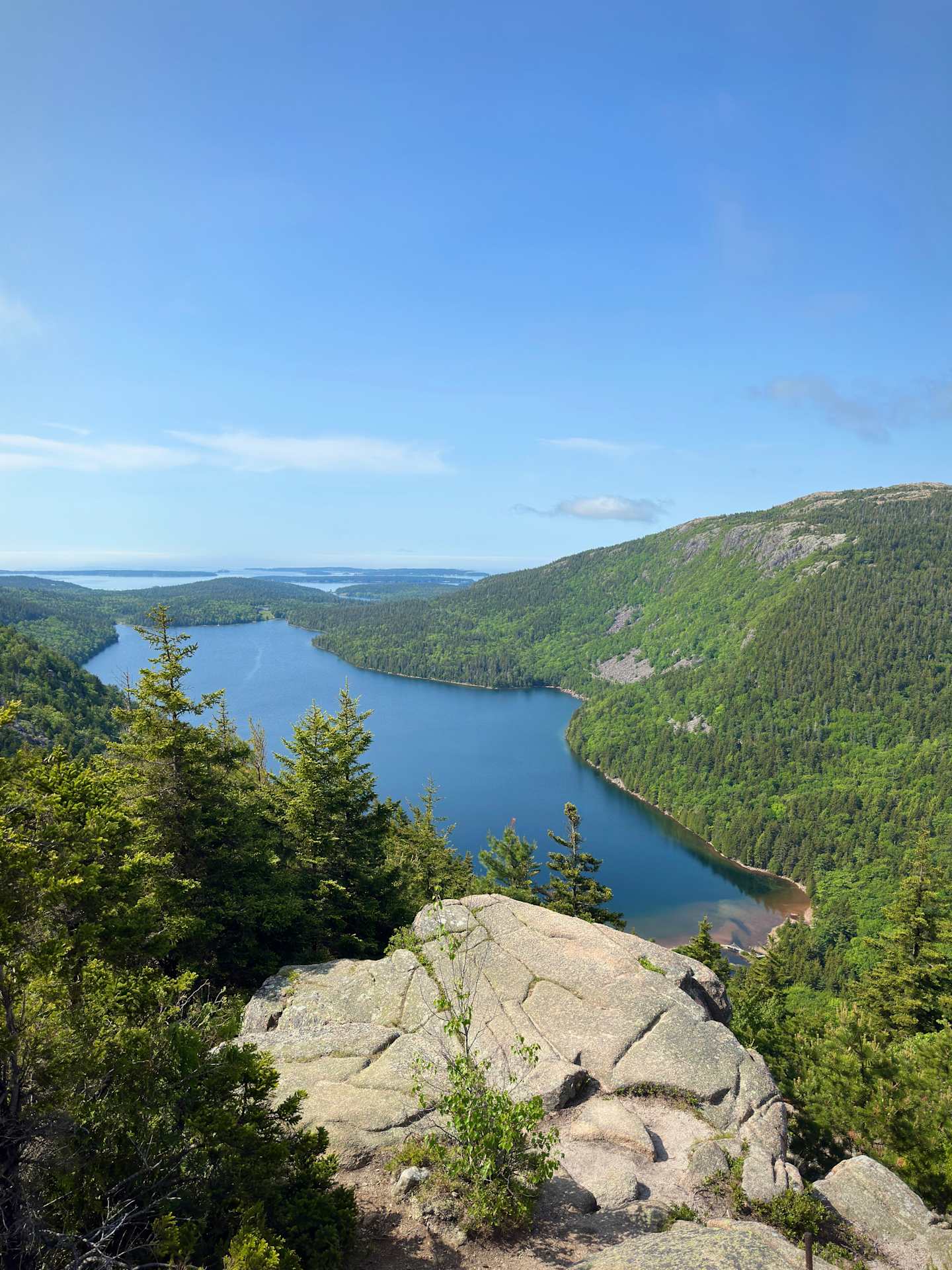 A scenic landscape with a rocky outcrop in the foreground overlooking a serene lake surrounded by lush, forested hills in the background under a clear blue sky.