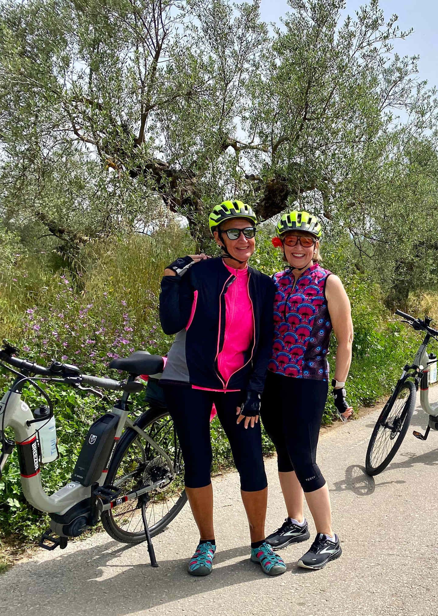Two women in cycling gear stand next to their bicycles in a scenic outdoor setting with lush greenery and trees in the background.