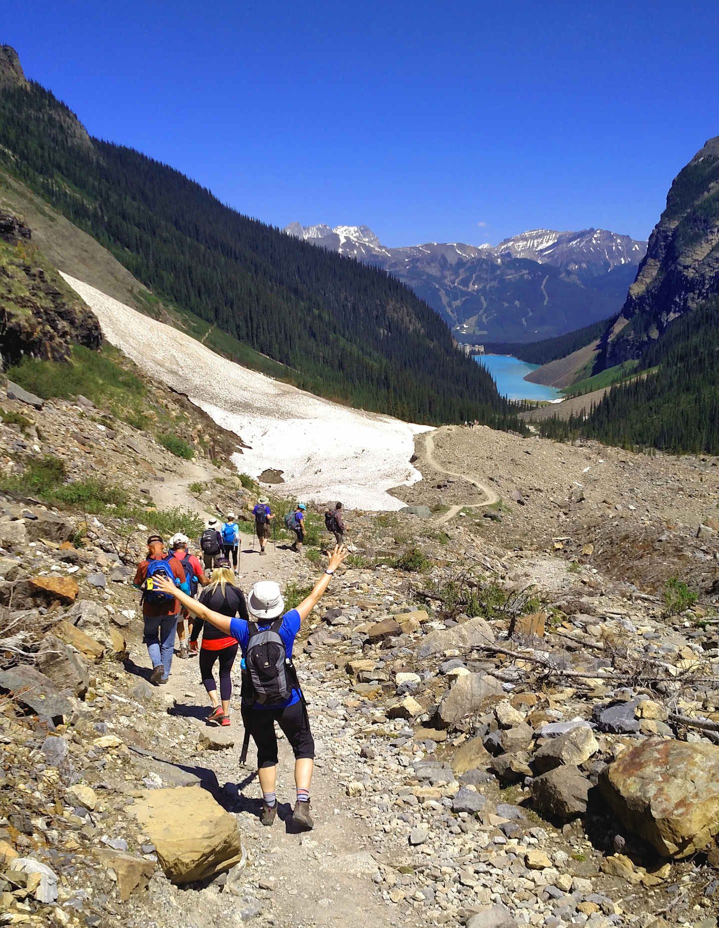 A group of hikers traversing a rocky trail in a mountainous landscape, with a stunning blue lake and snow-capped peaks visible in the background.
