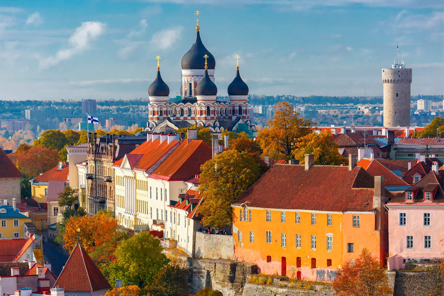 A vibrant cityscape with colorful buildings, a historic cathedral with onion-shaped domes, and a tower in the background, all set against a backdrop of a blue sky with fluffy clouds.