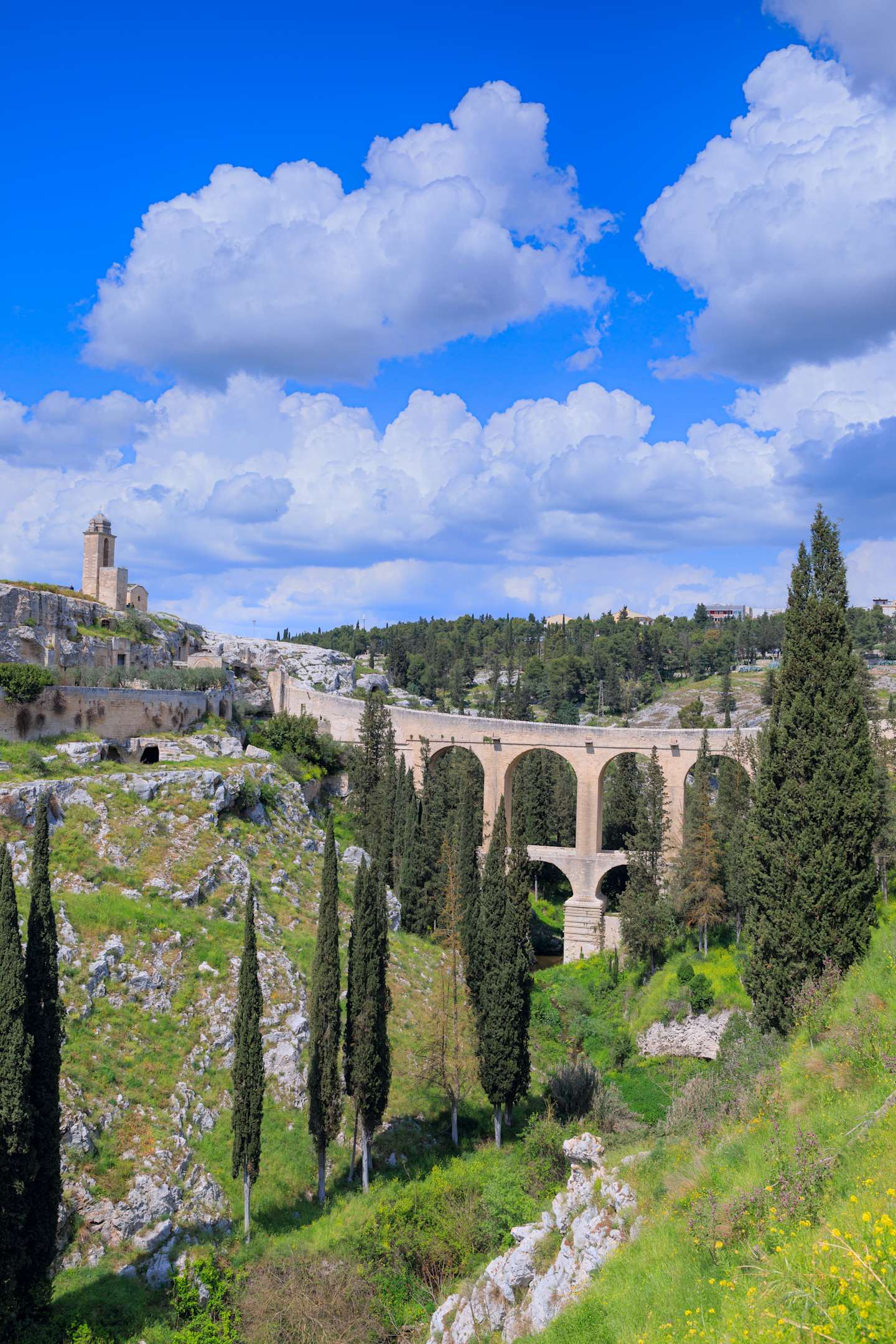A picturesque landscape featuring a grand stone bridge spanning a deep ravine, surrounded by lush greenery and a vibrant blue sky dotted with fluffy white clouds.