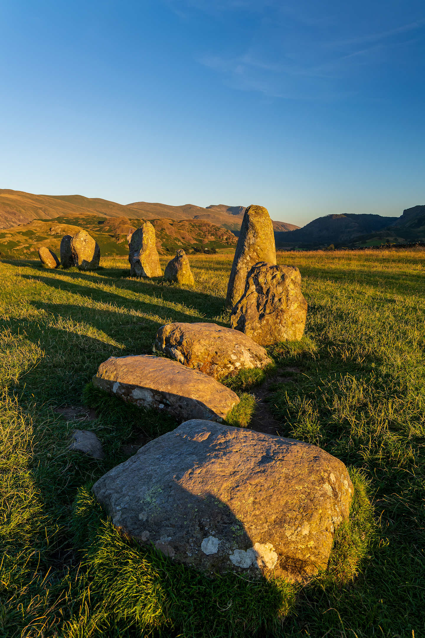 A field of large, weathered stone formations stands in the foreground, with rolling hills and a clear blue sky in the background.