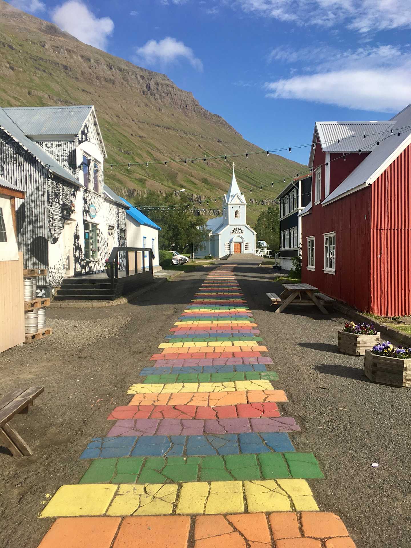 A colorful brick path leads through a quaint village nestled between rolling hills, with a white church steeple visible in the distance.
