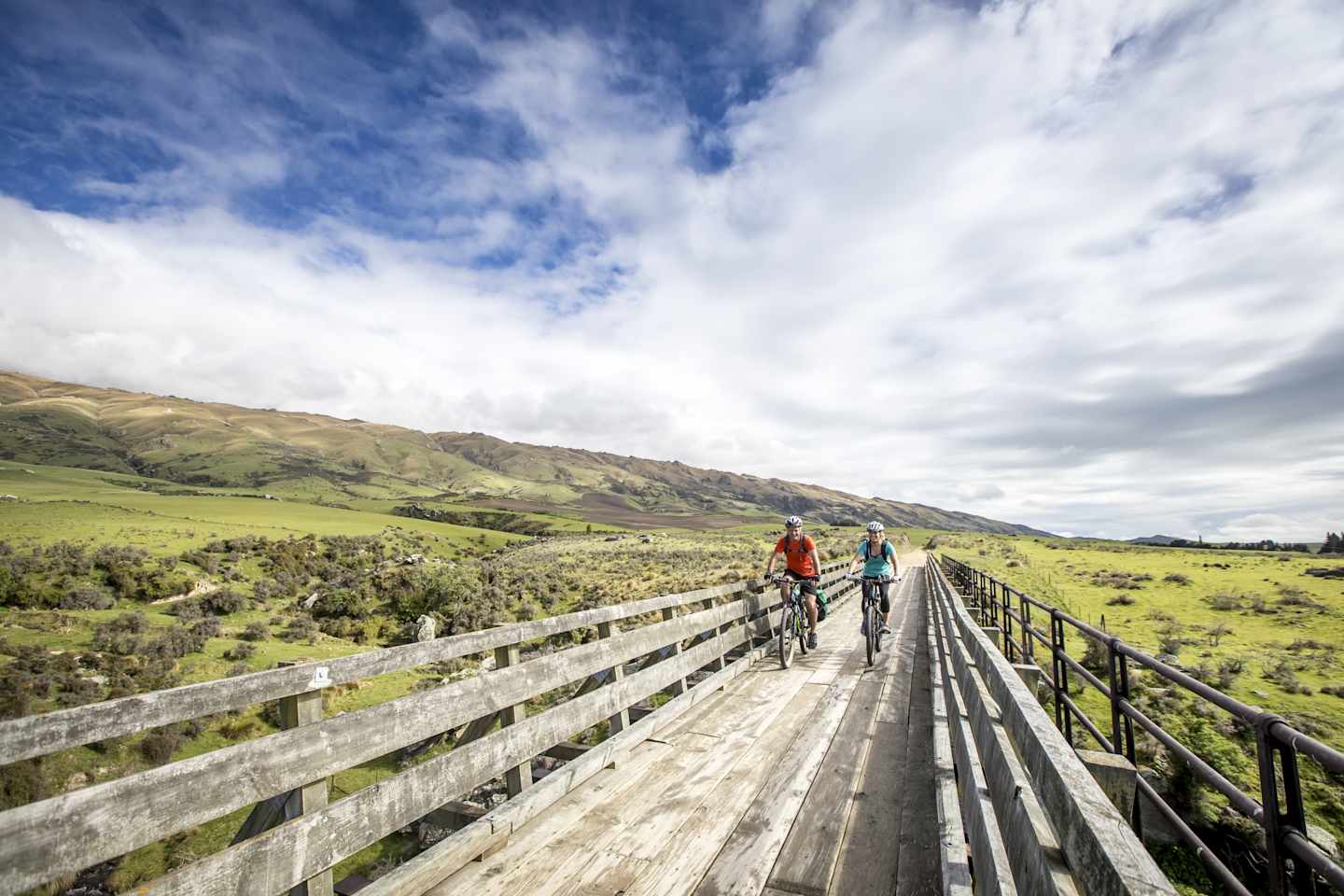 A wooden bridge traverses a grassy landscape, with two cyclists riding across it against a backdrop of rolling hills and a cloudy sky.