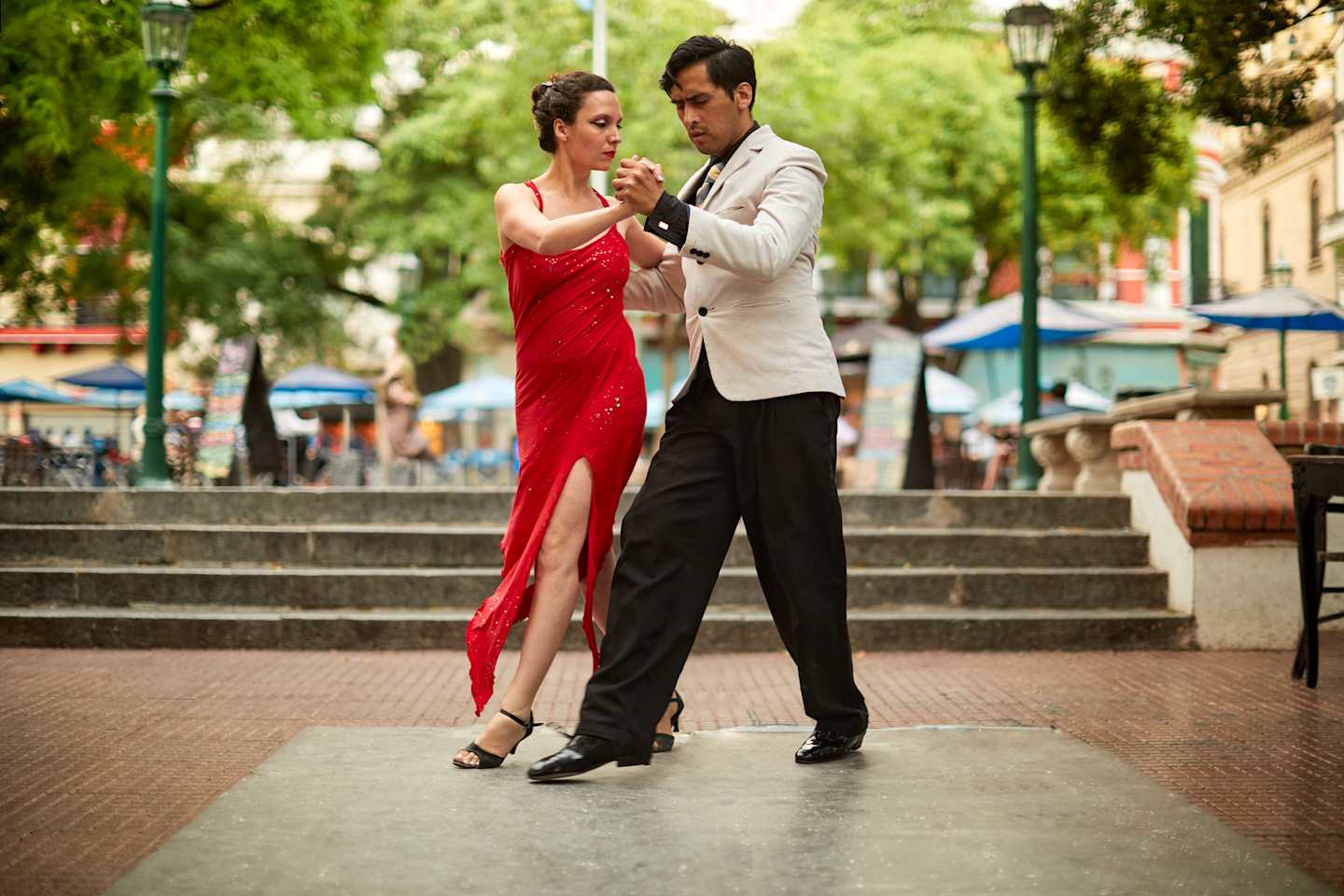 A couple dressed in formal attire are dancing on a set of stairs in an outdoor urban setting, surrounded by lush greenery and buildings.