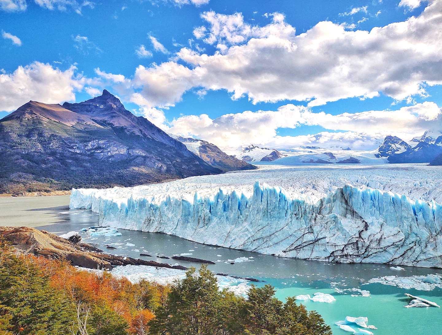 A majestic glacier with towering peaks in the background, surrounded by a serene turquoise lake and vibrant autumn foliage in the foreground.