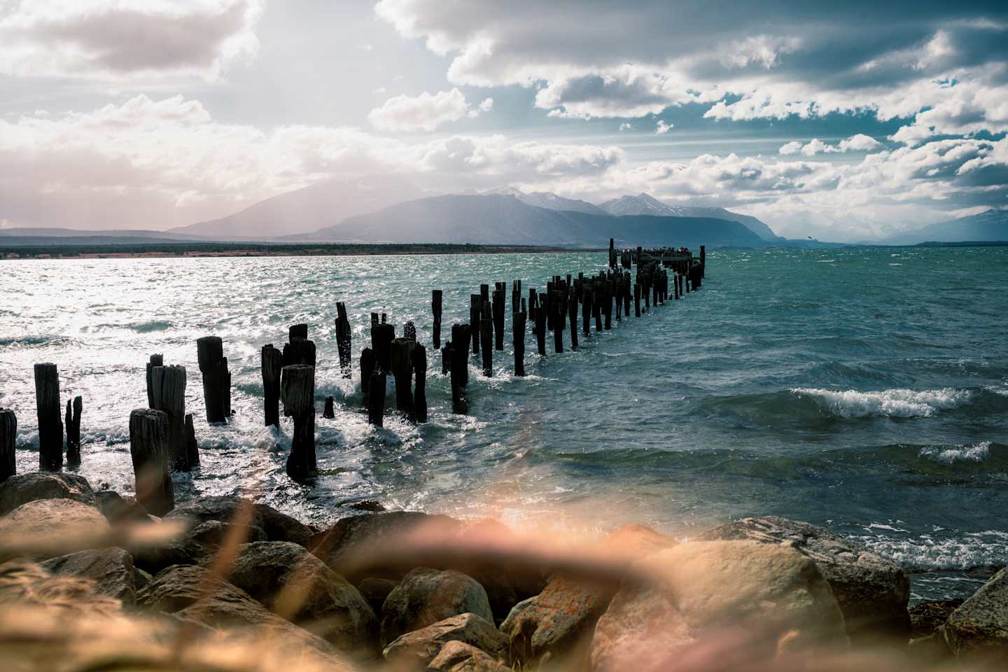 A rugged coastline with a wooden pier extending into the turbulent waters, set against a dramatic sky with billowing clouds, creating a captivating and moody landscape.
