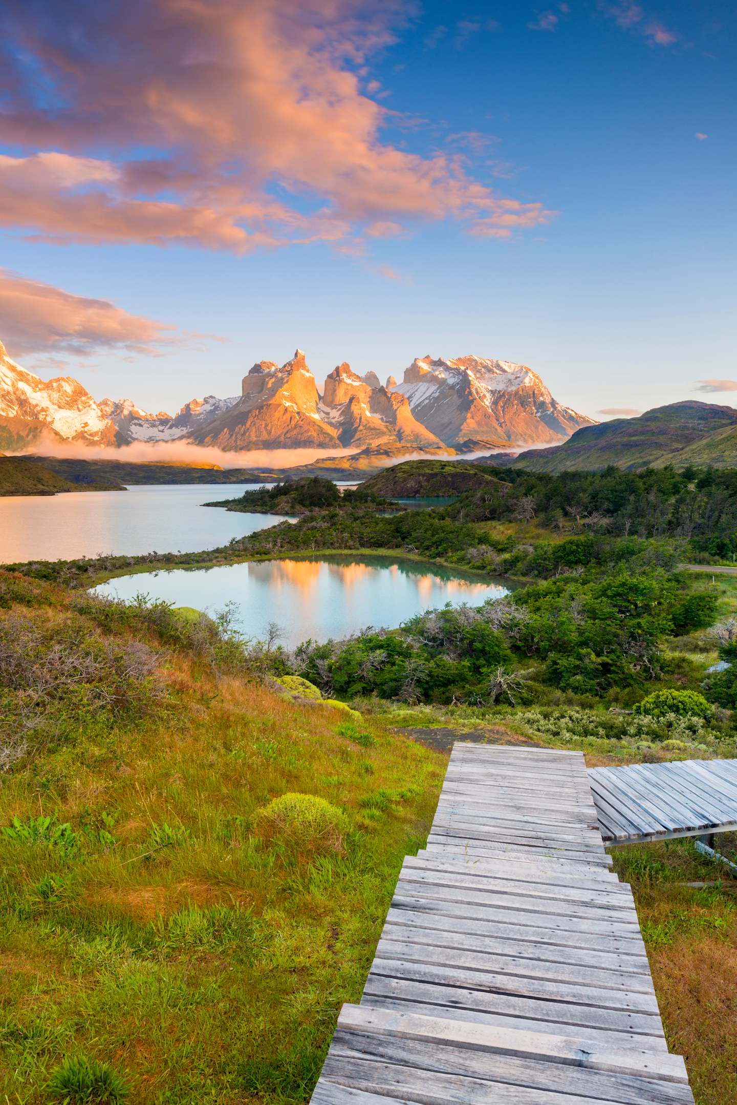 A wooden walkway leads through a lush, grassy landscape towards a serene lake, with majestic mountains bathed in the warm glow of the setting sun in the background.
