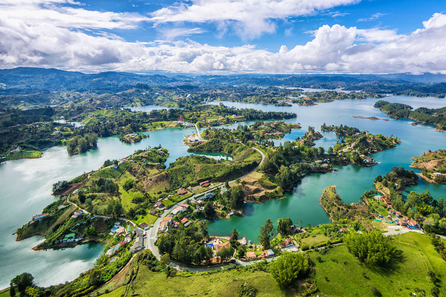 A breathtaking aerial view of a lush, green landscape dotted with islands and surrounded by crystal-clear, turquoise waters, all set against a backdrop of towering mountains and a vast, blue sky with fluffy white clouds.