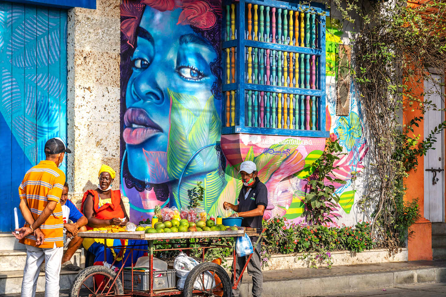 A vibrant and colorful mural adorns the wall, depicting a striking blue face with expressive features. In the foreground, a vendor sells fresh produce from a bicycle cart, adding to the lively and bustling atmosphere of the scene.