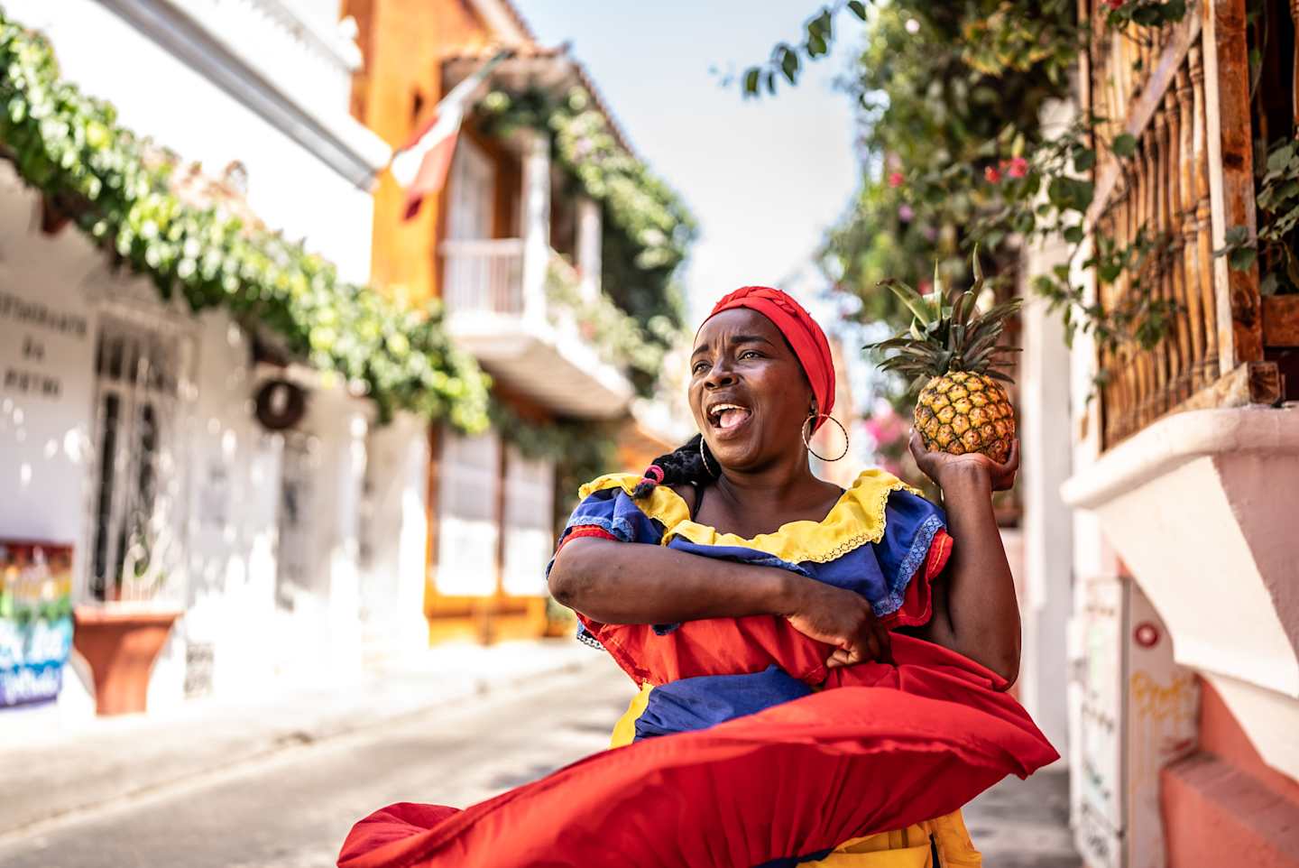 A vibrant and joyful woman in colorful traditional clothing stands on a street lined with lush greenery and buildings, holding a pineapple and smiling brightly.