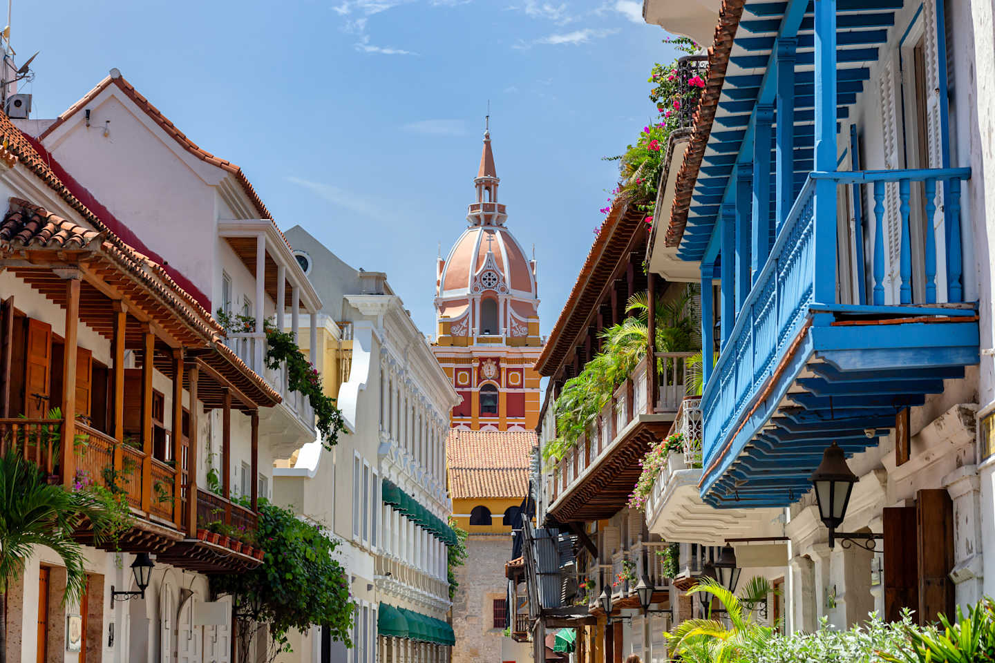 A colorful and vibrant street scene with traditional colonial-style buildings, balconies, and a prominent church tower in the background.