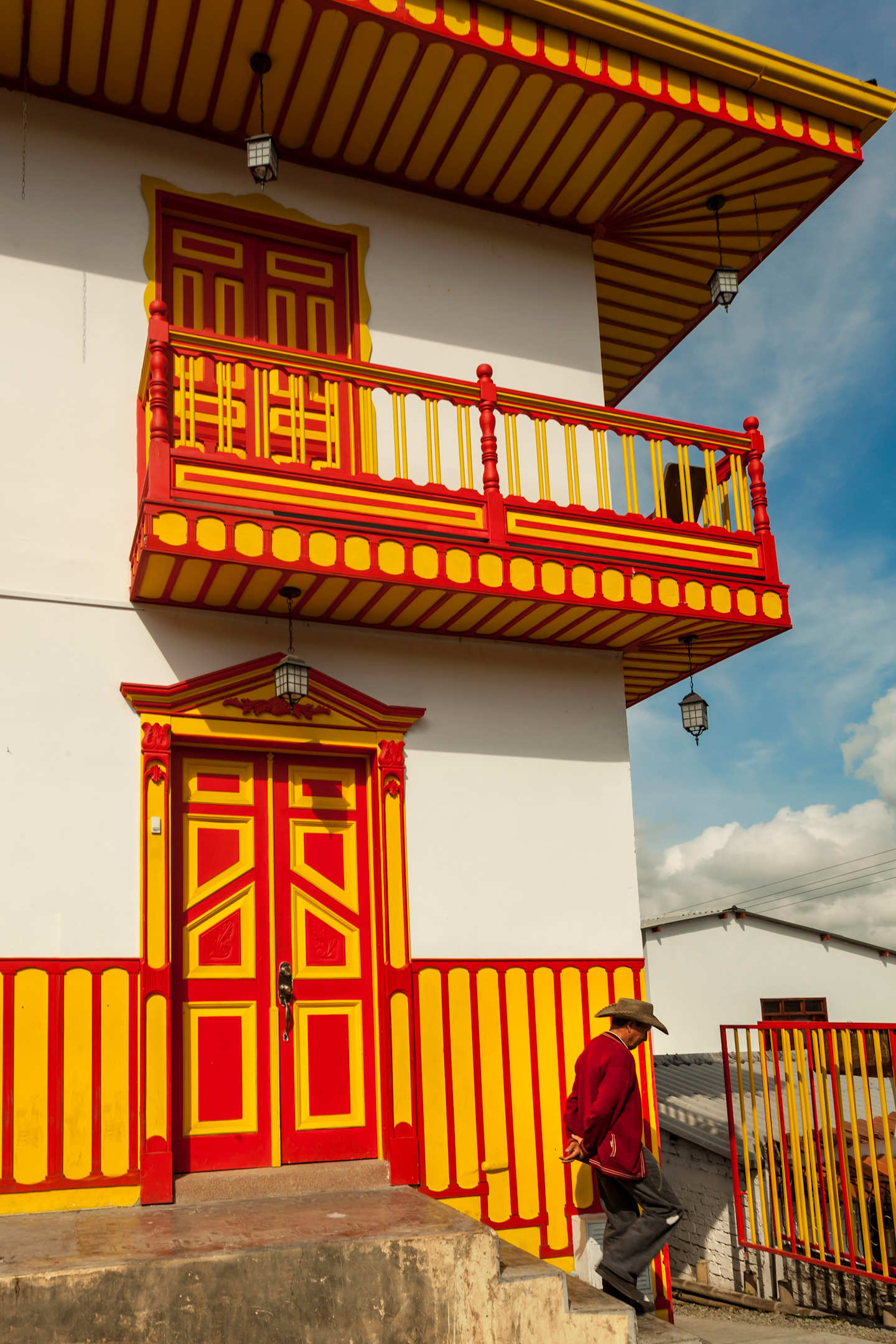 A colorful, traditional-style building with a balcony and ornate red and yellow decorations, set against a cloudy sky, with a person sitting on a bench in the foreground.