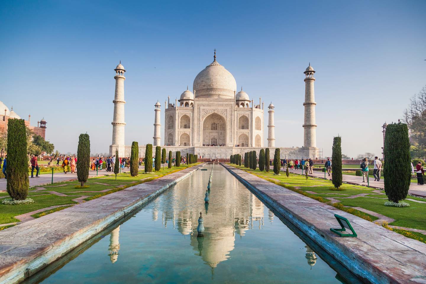 A magnificent white marble mausoleum, the Taj Mahal, stands majestically against a clear blue sky, reflected in a serene pool of water in the foreground, surrounded by lush greenery and symmetrical gardens.