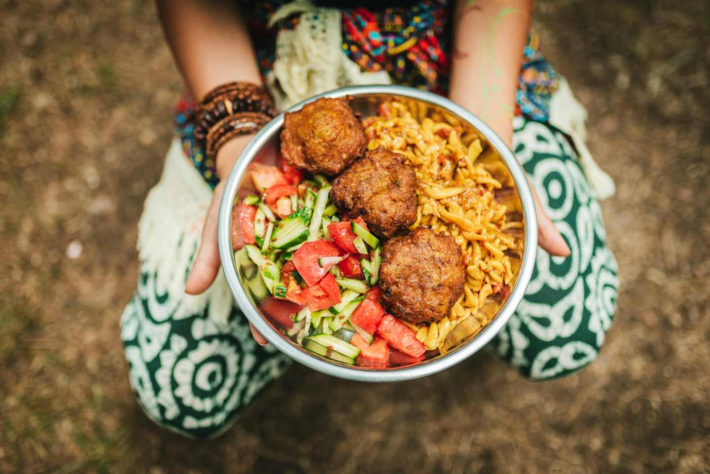 A plate of fried food, including what appears to be chicken or meat patties, sits in the hands of a person wearing a patterned dress against a grassy background.