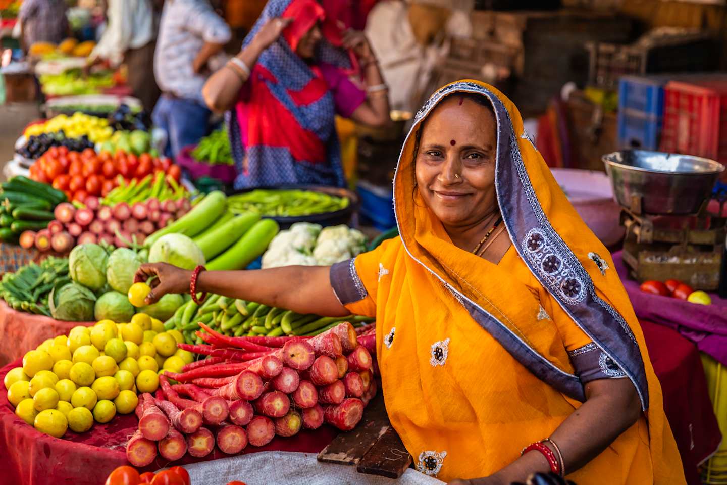 A vibrant outdoor market scene with a woman in a colorful yellow sari standing amidst an array of fresh produce, including various fruits and vegetables.