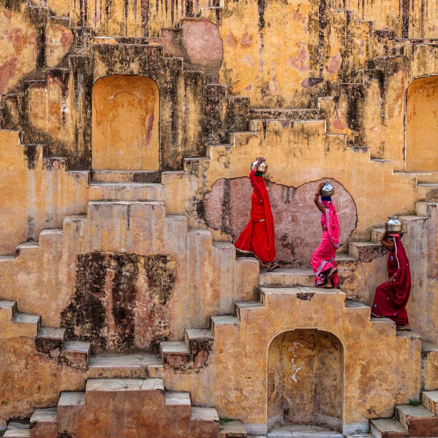 The image depicts two individuals in vibrant red robes ascending a set of ancient, weathered stone steps against a backdrop of crumbling, ochre-colored structures.