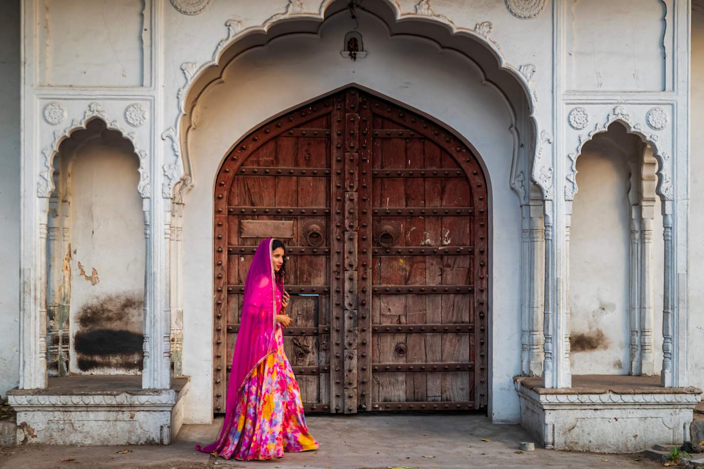 A vibrant, colorful dress stands in contrast to the ornate, weathered wooden doors and arched entryway of an architectural structure, creating a striking visual juxtaposition.