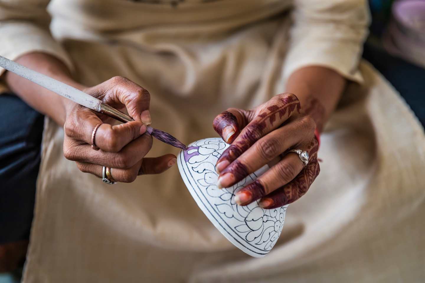 The image shows a person's hands decorating a white shoe with intricate patterns, against a neutral background.