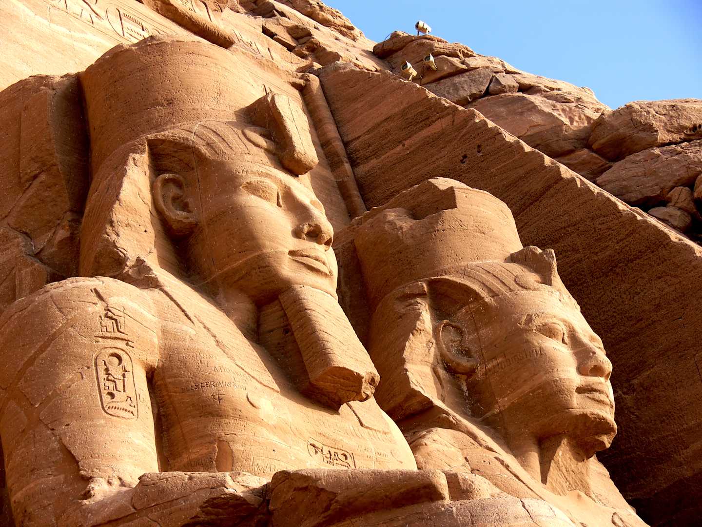 Massive carved stone statues of ancient Egyptian pharaohs against a backdrop of rocky cliffs under a bright blue sky.