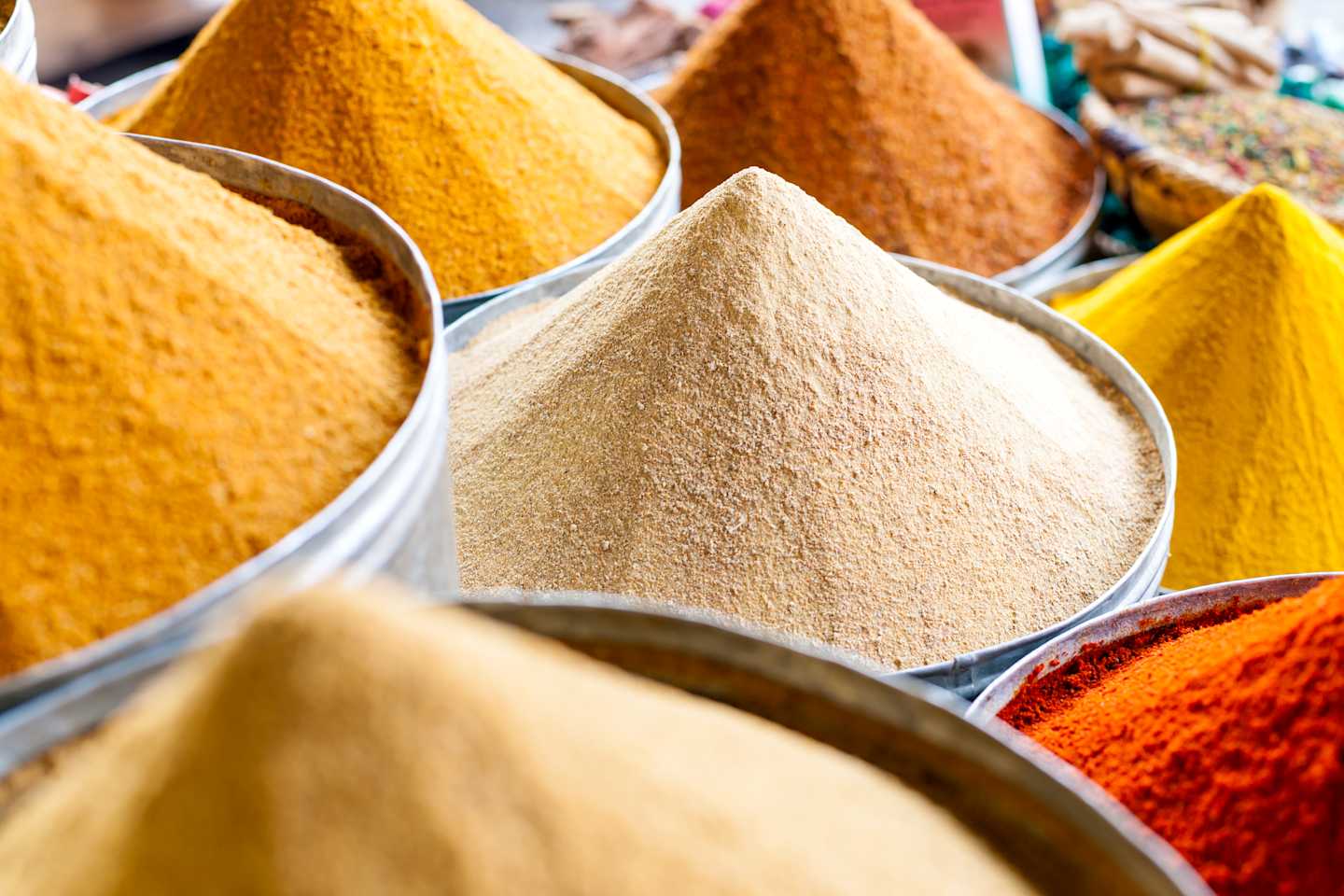 Vibrant piles of various spices and seasonings, including yellow, orange, and brown powders, fill the frame, creating a visually striking and textured display.