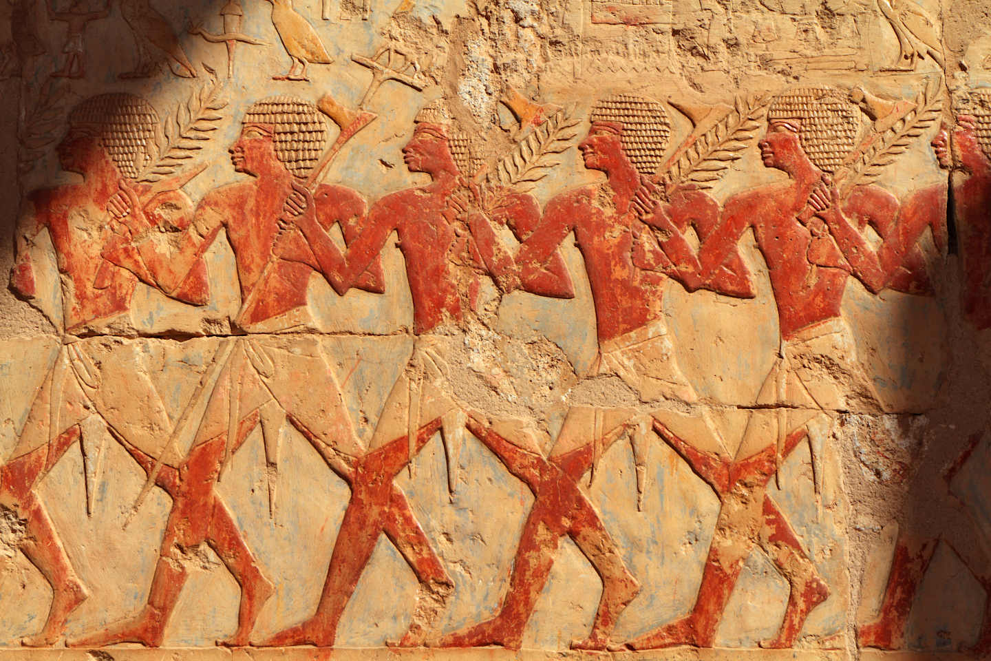 The image depicts a wall relief featuring a procession of stylized human figures in vibrant red and ochre tones, set against a textured stone background.