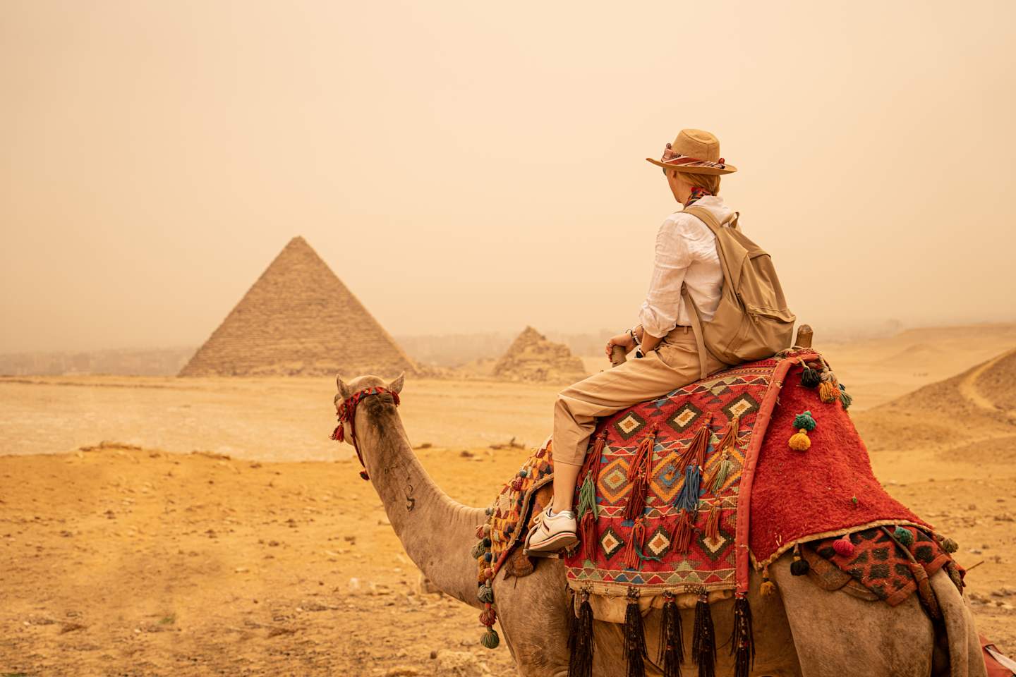 A person riding a camel in the foreground, with the iconic pyramids of Giza visible in the background against a hazy, desert landscape.