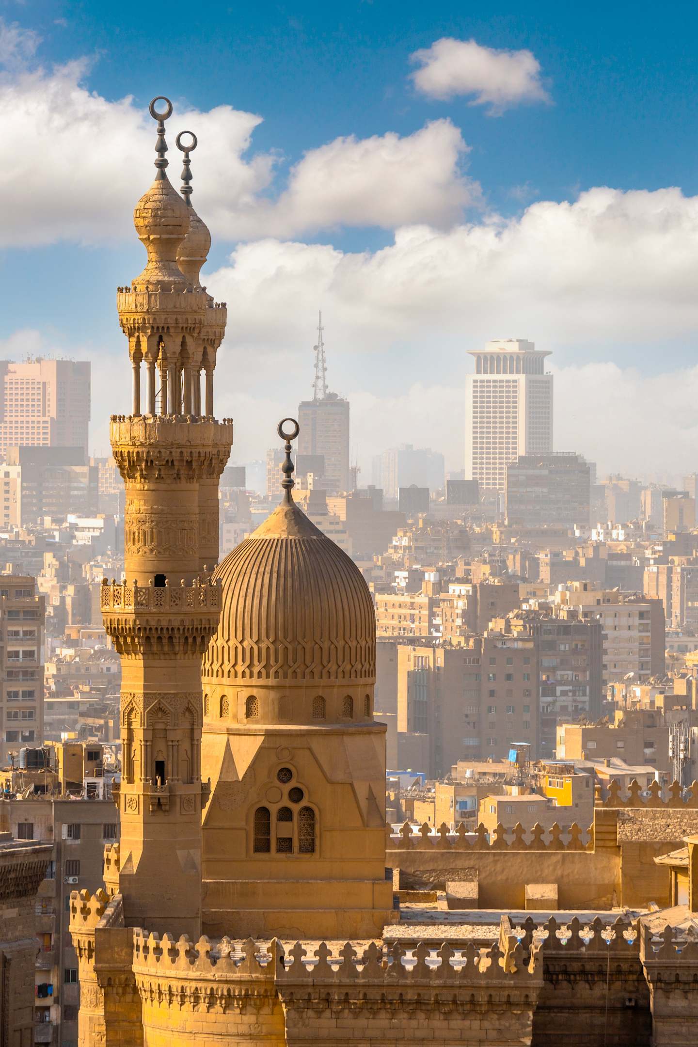 A majestic golden-domed mosque stands prominently in the foreground, surrounded by a bustling cityscape of high-rise buildings in the background under a bright, cloudy sky.