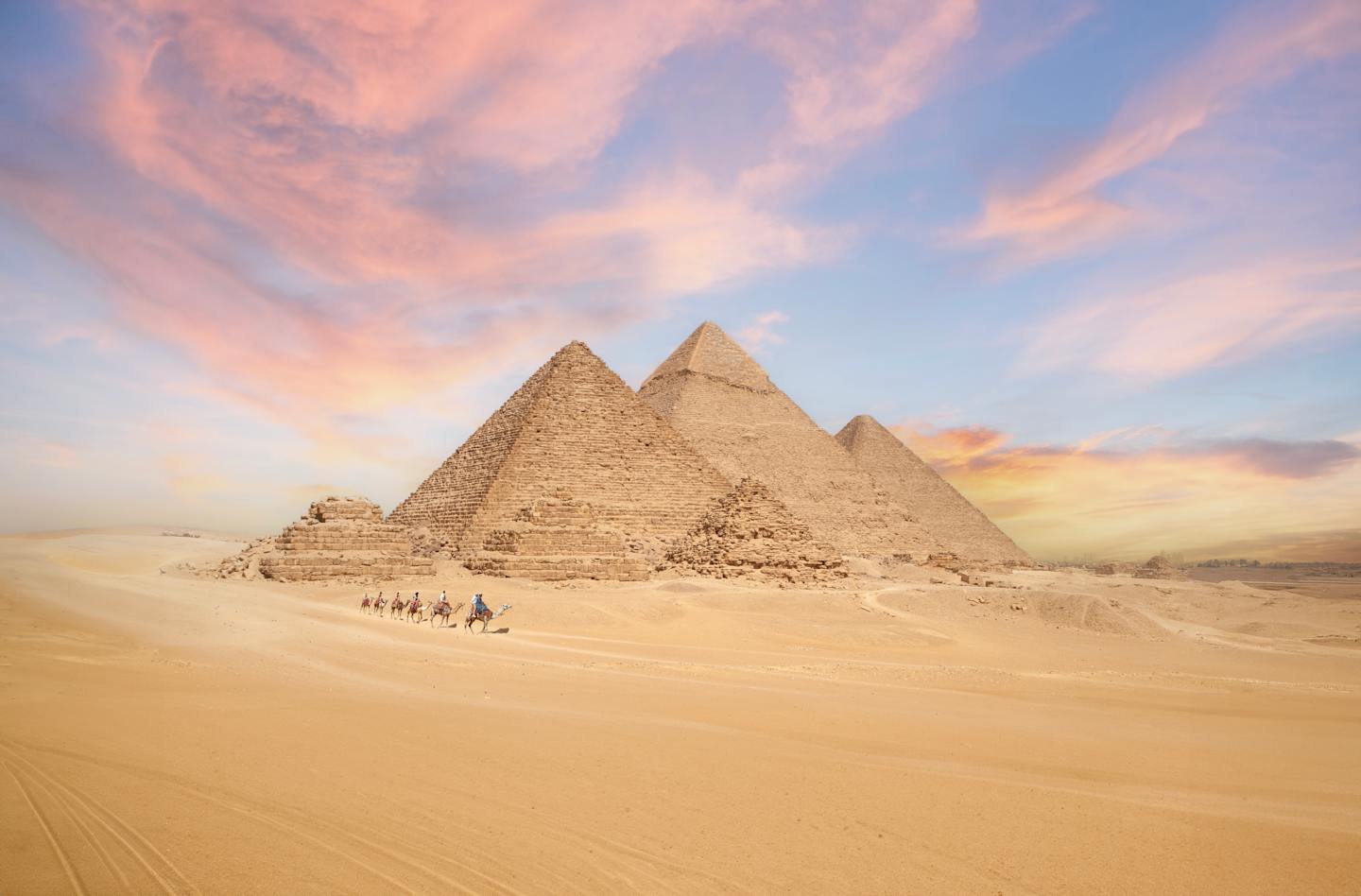 The image depicts the iconic Pyramids of Giza in Egypt, with their distinctive triangular shapes silhouetted against a vibrant, colorful sky at sunset. The vast expanse of desert sand in the foreground leads the viewer's eye towards the ancient monuments, creating a striking and awe-inspiring scene.