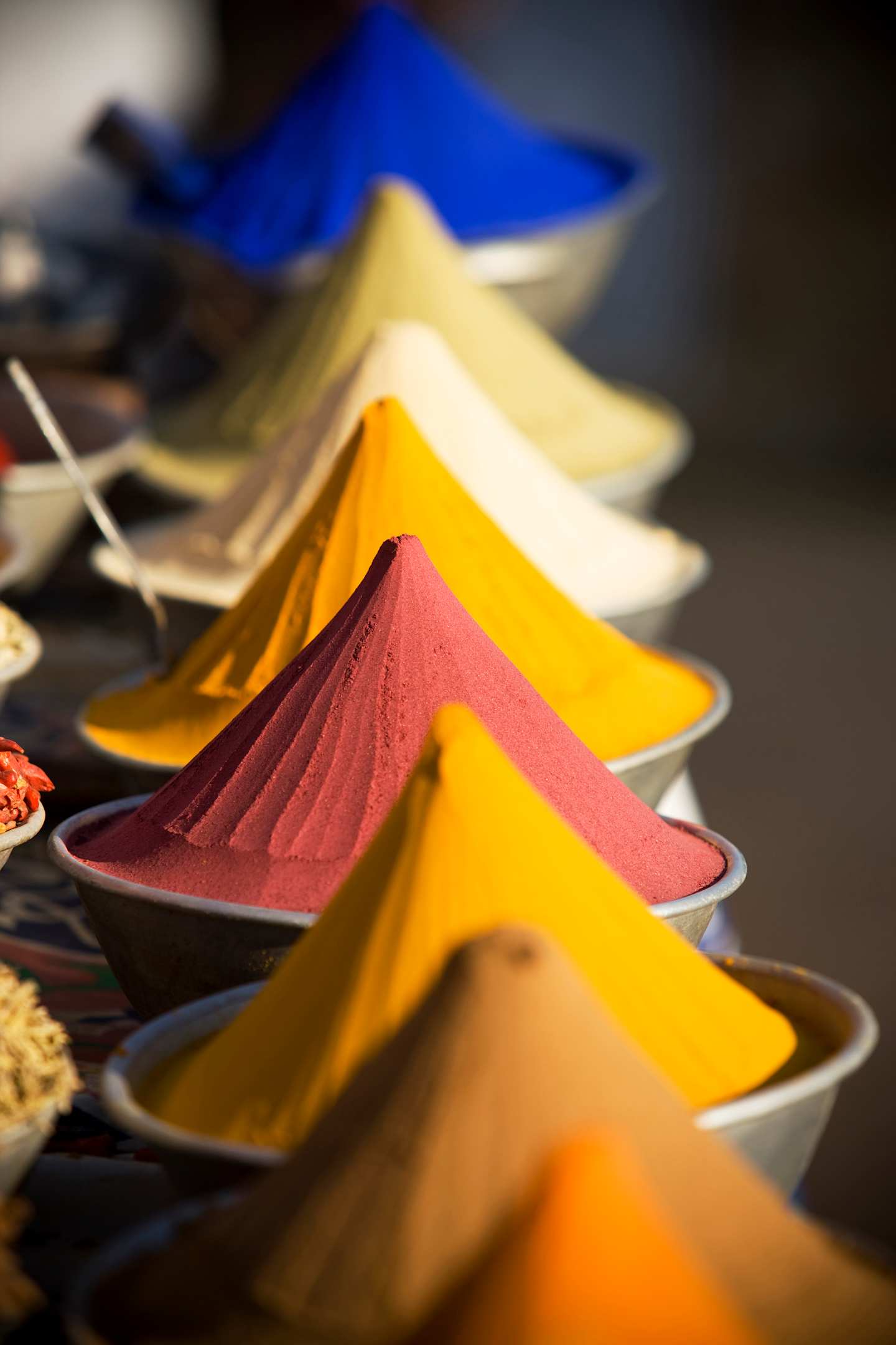 Vibrant, triangular-shaped spice piles in various colors, including blue, yellow, and red, are arranged in the foreground against a dark background.