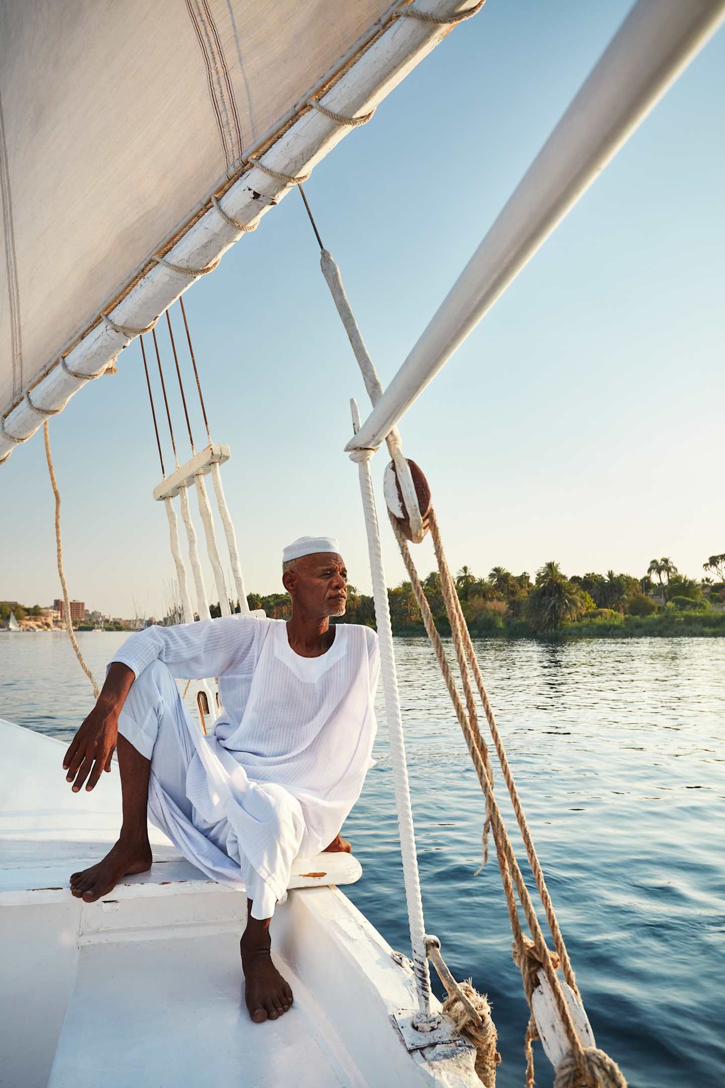 A person in a white robe sits on the deck of a sailboat, with the Nile River and lush greenery visible in the background.