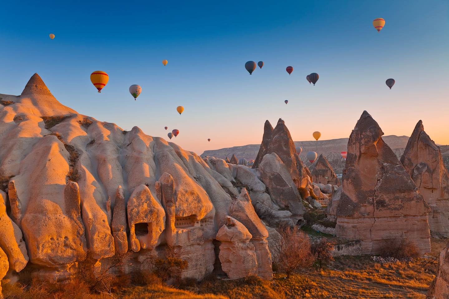 Colorful hot air balloons float above the unique rock formations of Cappadocia, Turkey, set against a vibrant blue sky at sunrise.