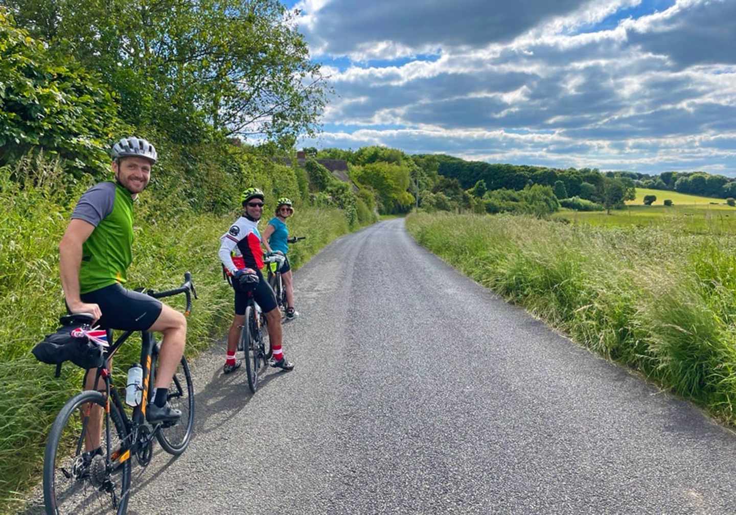 A group of cyclists riding on a gravel path surrounded by lush greenery and a scenic countryside landscape under a cloudy sky.