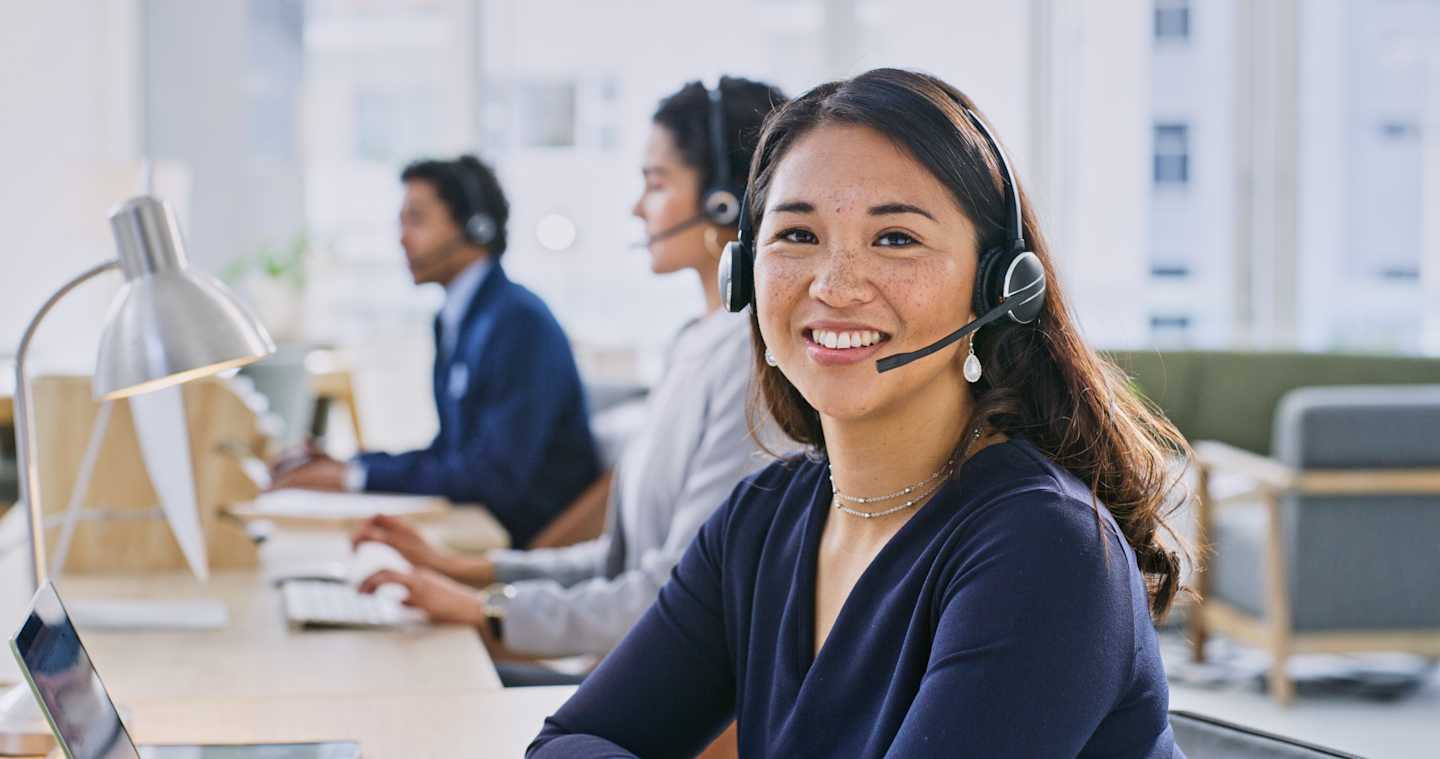 A smiling woman wearing a headset sits at a desk in an office setting, with other employees visible in the background.