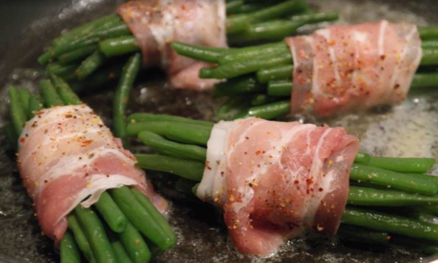 The image shows a close-up of green beans wrapped in slices of bacon, with a dark background suggesting a cooking surface or pan.