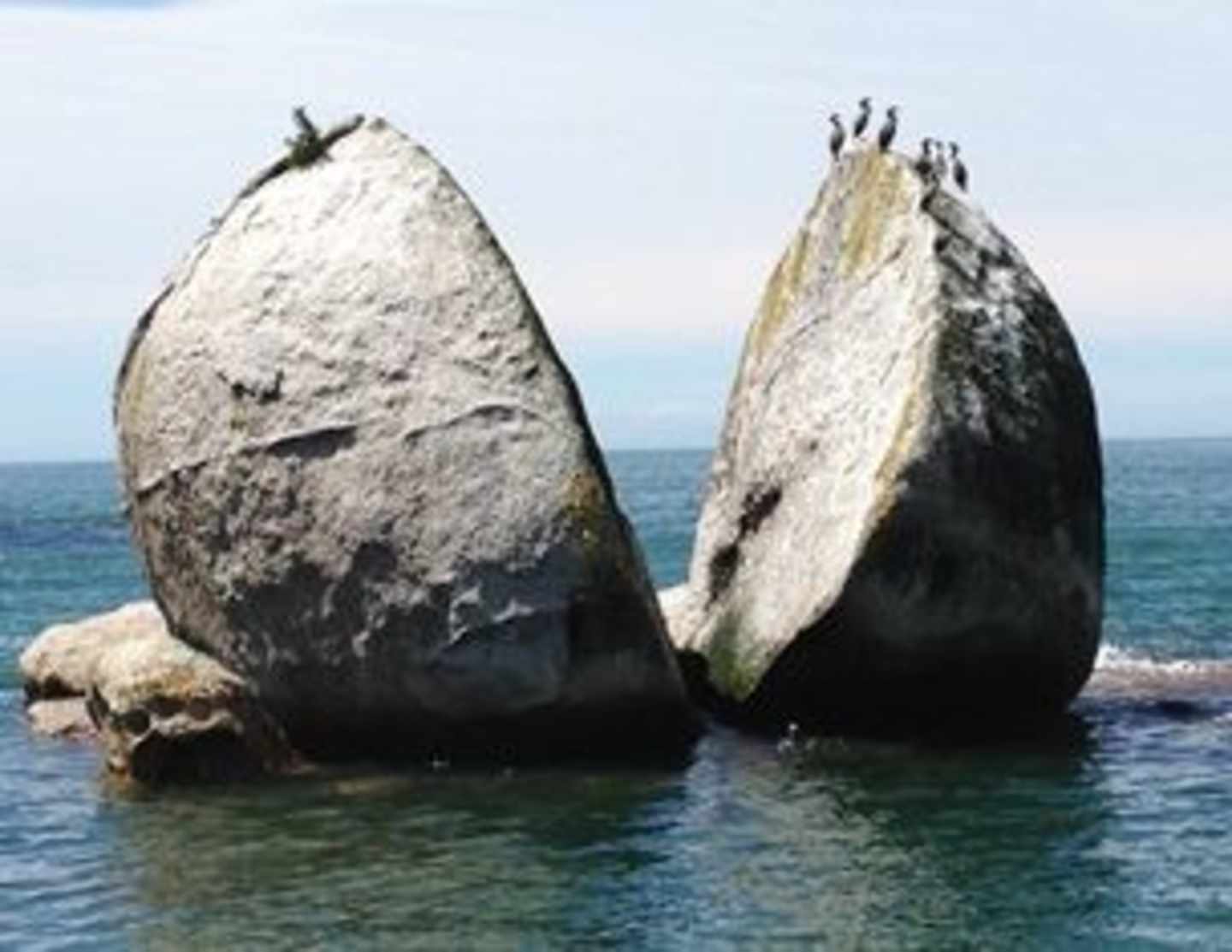 Two large, weathered rock formations emerge from the calm, turquoise waters of the ocean, with a clear blue sky in the background.