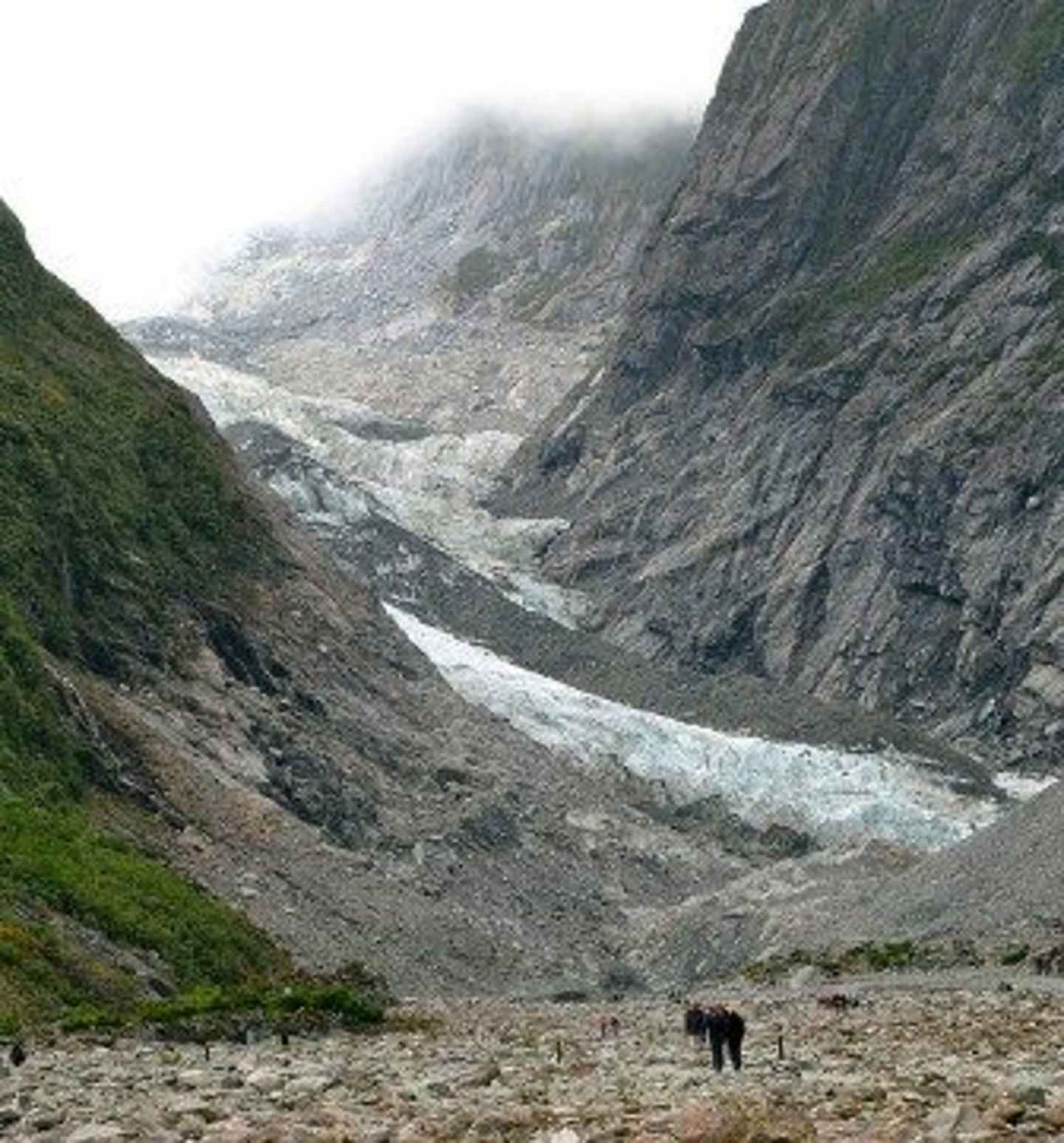 A rugged, mountainous landscape with a glacier-filled valley in the background, and a lone figure standing in the foreground against the dramatic scenery.