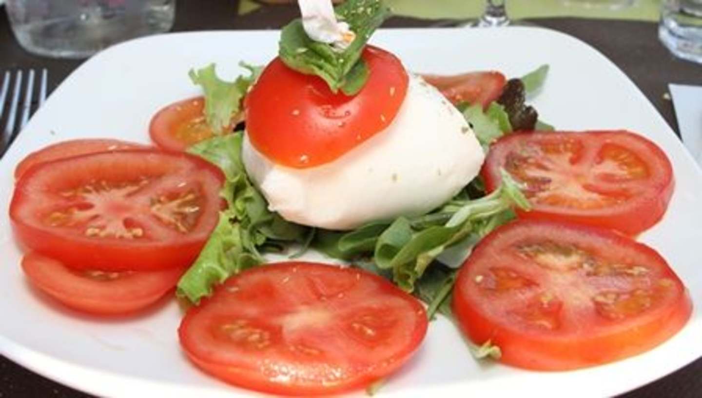 A white plate filled with fresh, vibrant tomato slices, a mozzarella cheese ball, and a green salad, set against a dark background.