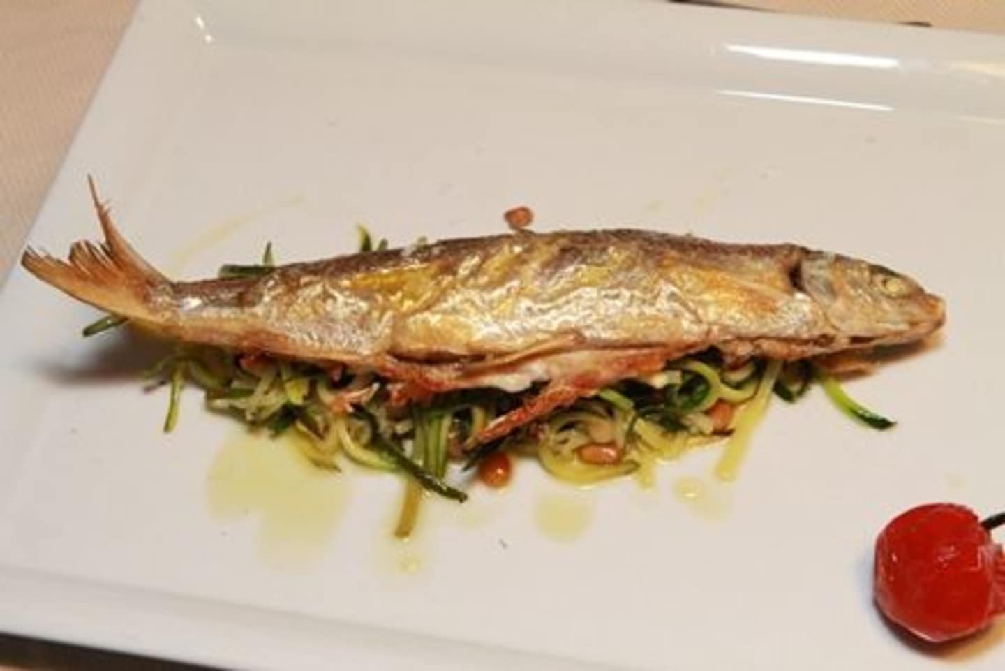 A grilled fish fillet rests on a bed of mixed greens, accompanied by a red chili pepper on a white plate.