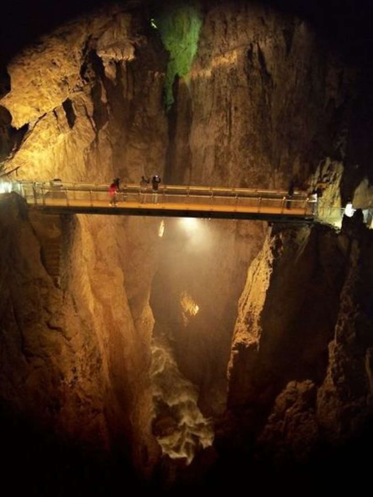 A dimly lit underground cave with a wooden bridge spanning across a deep chasm, surrounded by towering rock formations and stalactites.