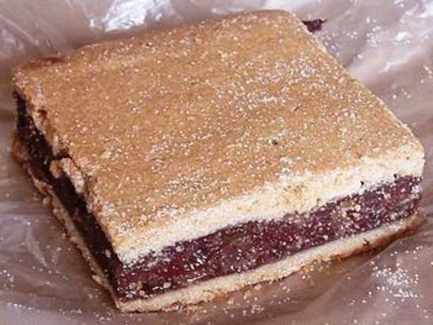 A square-shaped dessert with a golden-brown top and a dark filling, resting on a clear plastic surface.