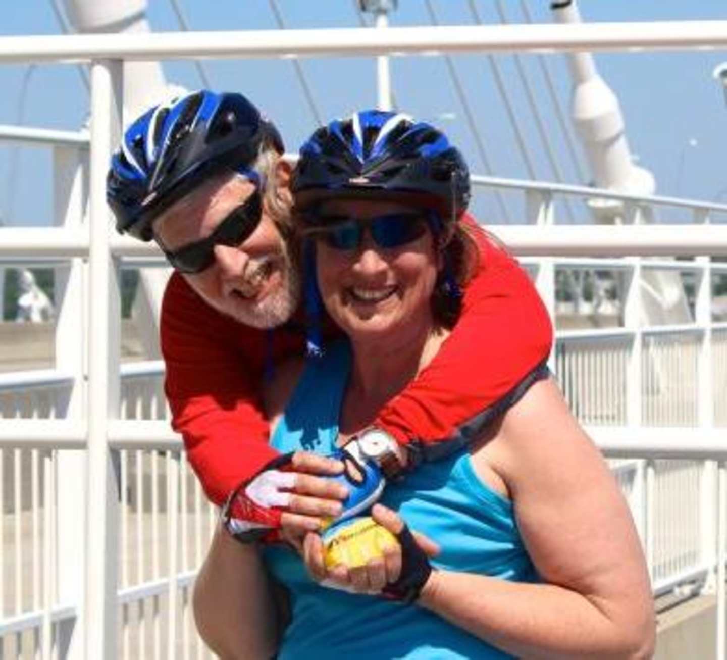 Two people wearing bicycle helmets and brightly colored athletic clothing are embracing each other in the foreground, with a metal railing and a blurred background visible behind them.