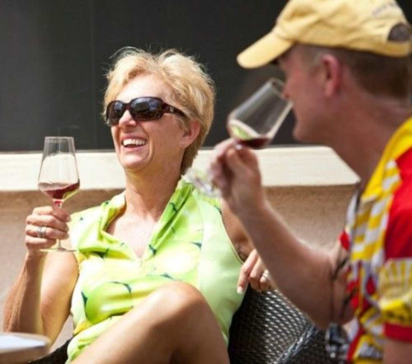A smiling woman in a bright green top is enjoying a glass of wine while conversing with a man wearing a colorful shirt in what appears to be a casual dining setting.