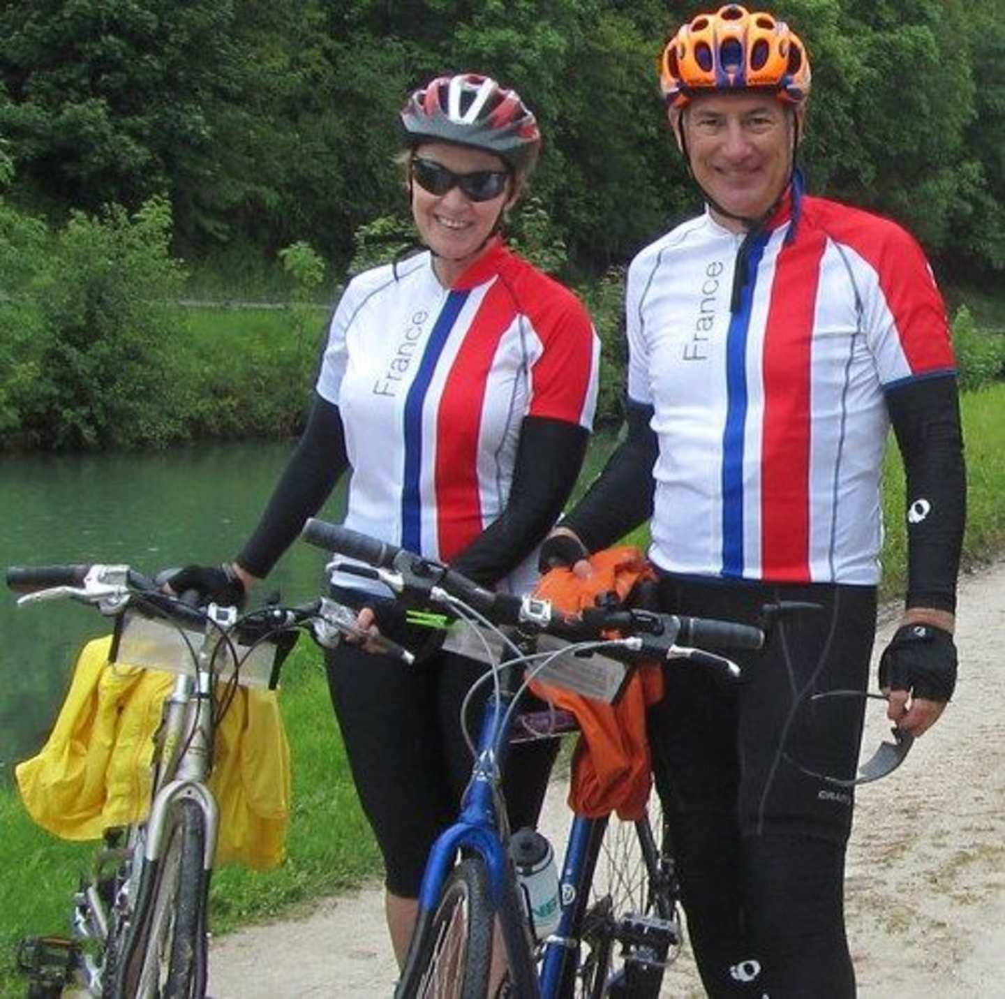 Two cyclists, a man and a woman, are standing with their bicycles in a lush, green outdoor setting with trees and a body of water visible in the background.