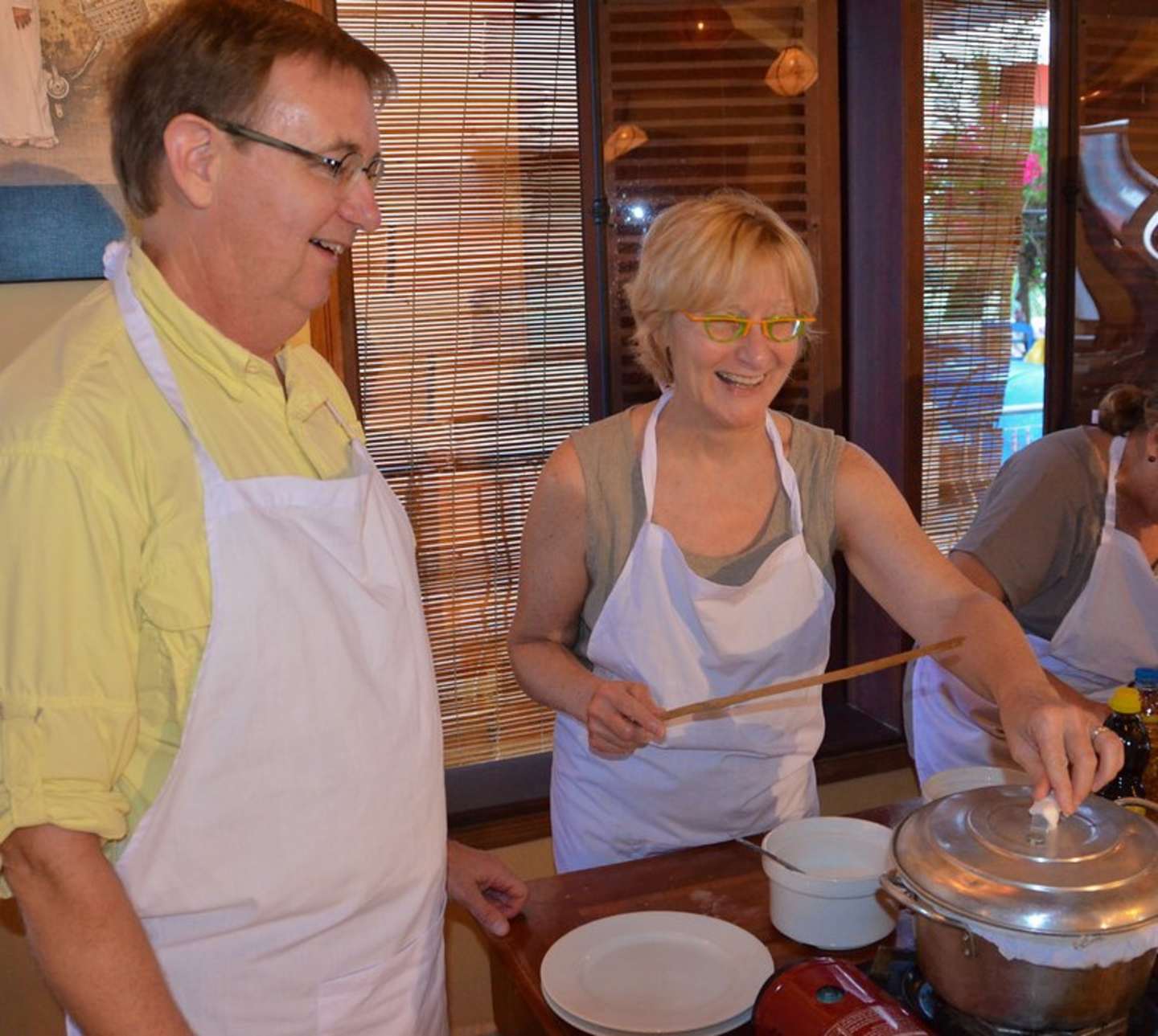 Two people, a man and a woman, are standing in a kitchen setting, wearing aprons and appearing to be engaged in some cooking or food preparation activity. The background includes a window with blinds and what appears to be a restaurant or cafe-like environment.
