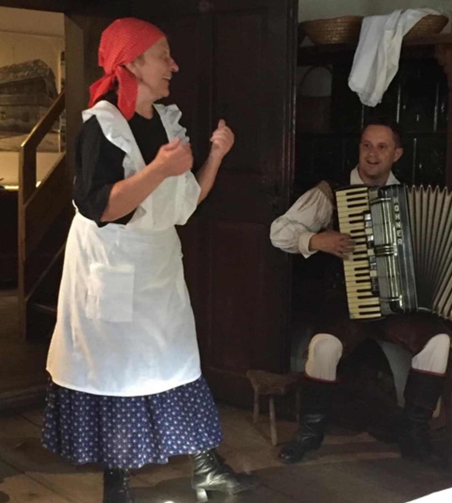 A woman in traditional folk costume, wearing a red headscarf and white apron, appears to be singing or performing, while a man plays an accordion in the background.
