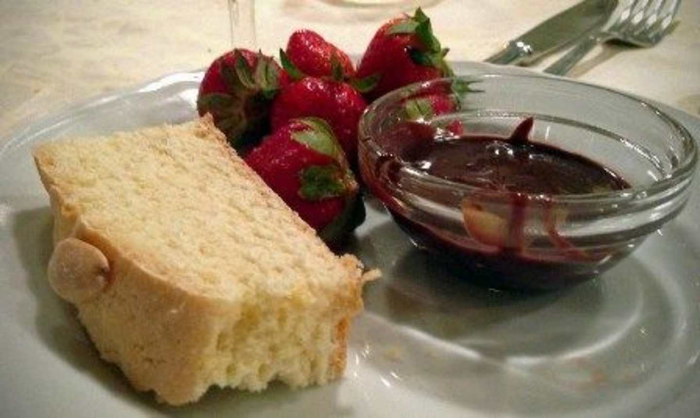 Fresh, ripe strawberries are arranged on a plate, accompanied by a slice of bread and a small glass container filled with a dark, viscous substance, likely a chocolate or fruit-based dipping sauce.