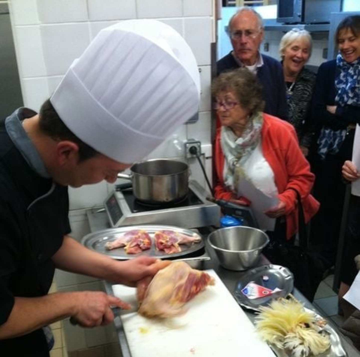 A chef in a white uniform is preparing food on a counter, surrounded by a group of older adults observing and interacting with the cooking process.