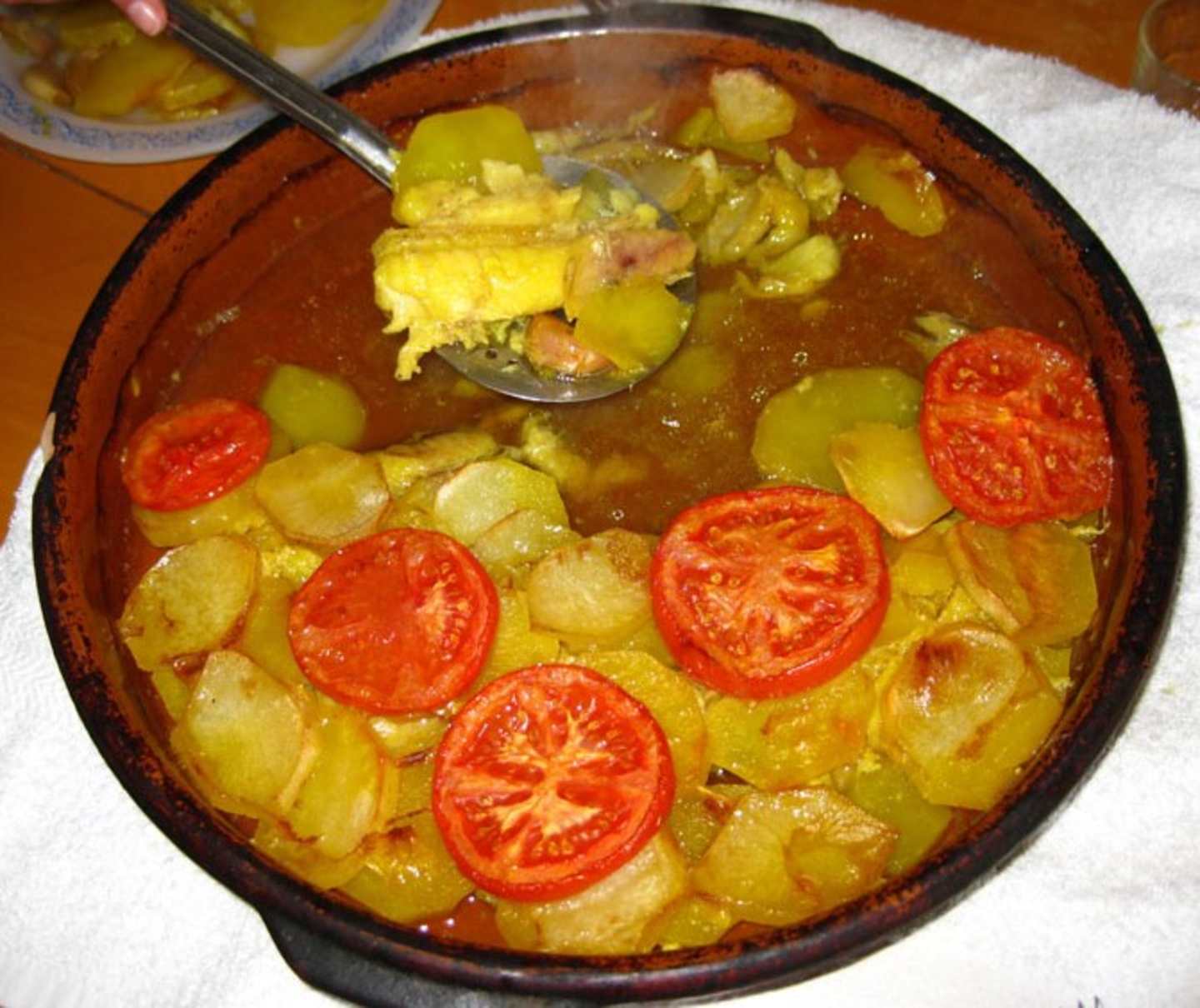 A bowl filled with a savory stew or soup, containing chunks of potatoes, tomato slices, and other vegetables, with a spoon visible in the foreground.